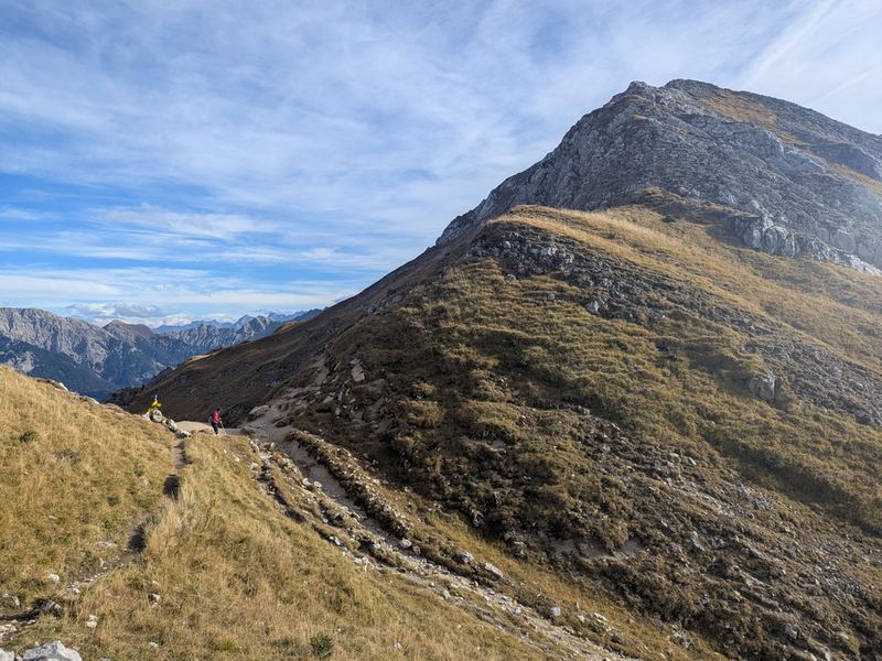 Grasflanke mit Pfad unter einem felsigen Gipfel, zwei Wanderer auf dem Weg